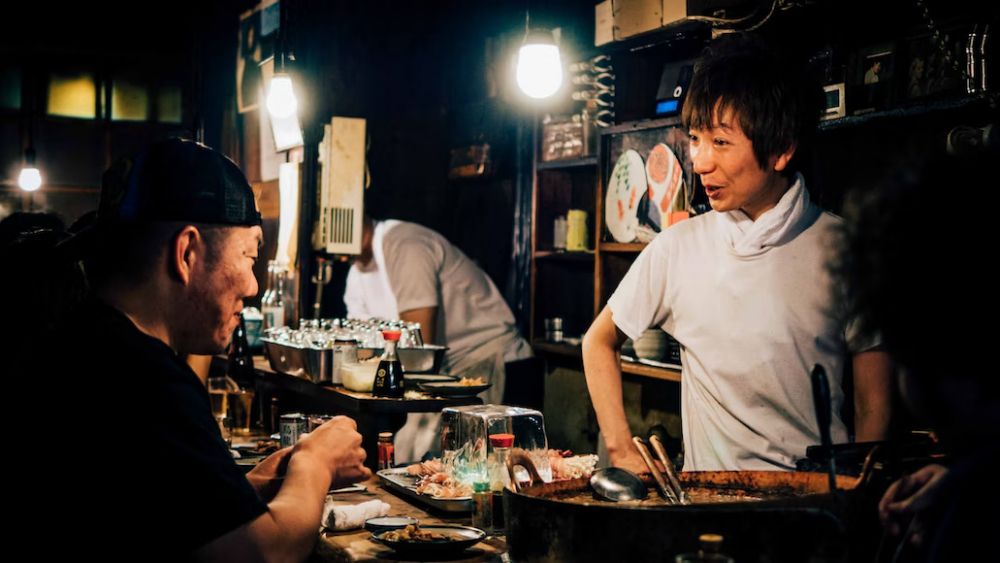 A Japanese food bar with a chef stood behind the bar talking to a smiling customer